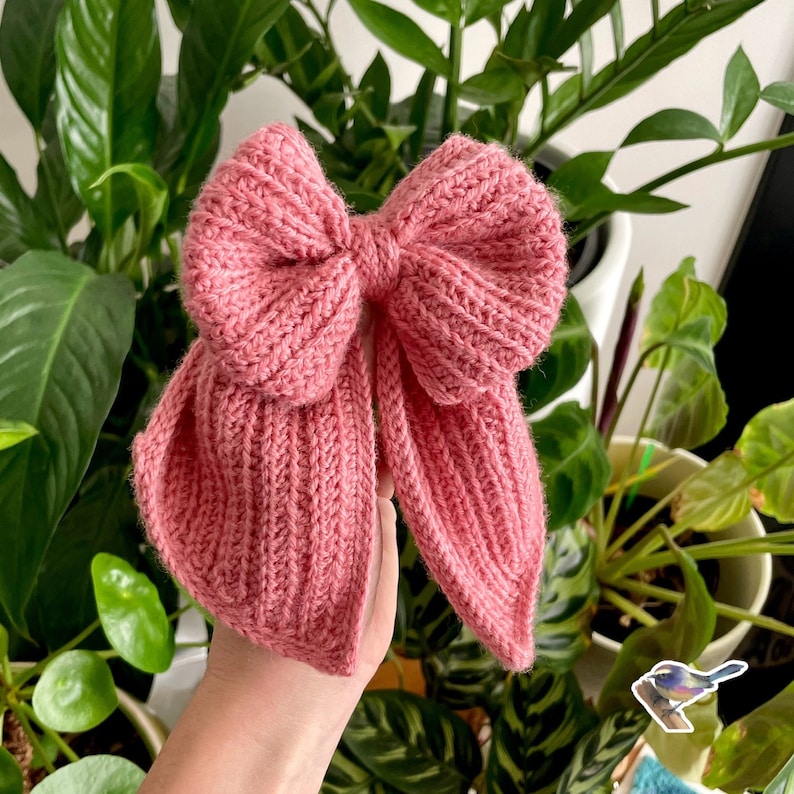 May include: A pink knitted bow with a textured surface. The bow is tied in a large knot and has long tails.
