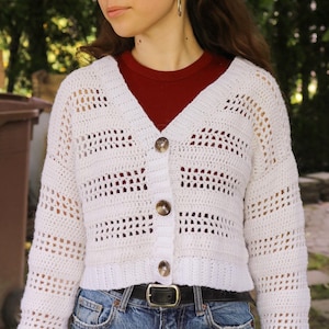 May include: A white, open-weave crochet cardigan with a V-neck and three brown buttons. The long-sleeved sweater is paired with blue jeans and a black belt. The cardigan's texture and design are prominent.