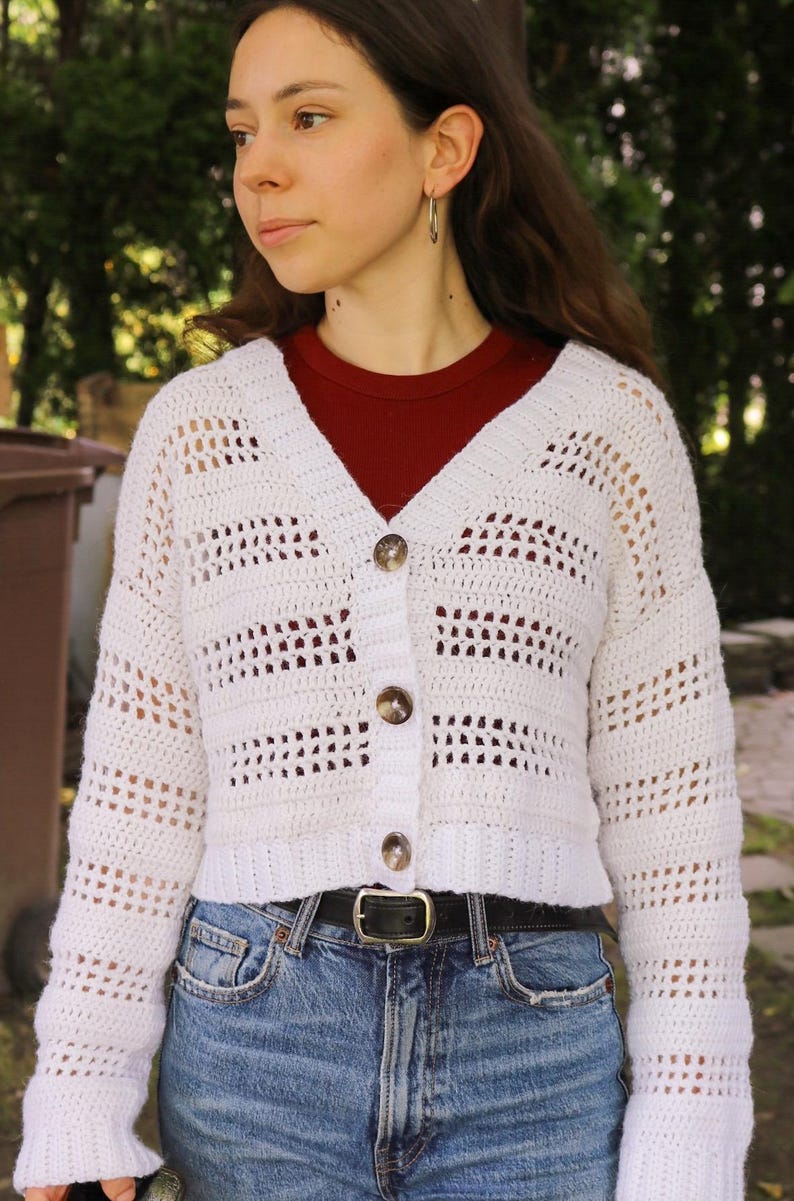 May include: A white, open-weave crochet cardigan with a V-neck and three brown buttons. The long-sleeved sweater is paired with blue jeans and a black belt. The cardigan's texture and design are prominent.