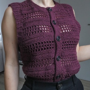 Puede incluir: Un chaleco sin mangas de ganchillo con cierre de botones. El chaleco es de color burdeos oscuro y tiene un delicado patrón calado.