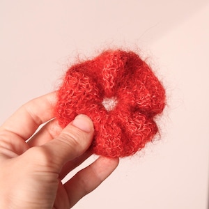 Könnte beinhalten: Ein leuchtend rotes Haargummi aus flauschigem, strukturiertem Material. Das Haaraccessoire ist kreisförmig mit einem Loch in der Mitte. Das Haargummi wird vor einem hellrosa Hintergrund gehalten.