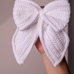May include: A white crocheted bow with long tails. The bow is tied in a knot and the tails are hanging down.