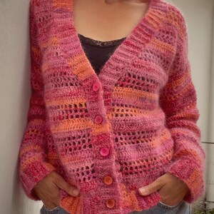 May include: A pink and orange knit cardigan with a button-up front. The sweater has a V-neck and a textured, open-weave design. The buttons are a matching pink color. The person is wearing blue jeans.