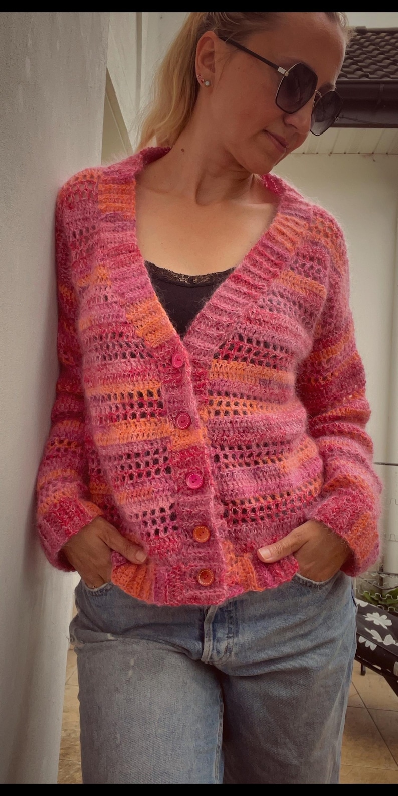 May include: A pink and orange knit cardigan with a button-up front. The sweater has a V-neck and a textured, open-weave design. The buttons are a matching pink color. The person is wearing blue jeans.