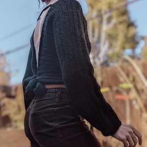 May include: A dark green, open-back sweater with long sleeves, tied at the waist. The sweater is paired with dark brown corduroy pants. The person is wearing glasses and has their hair styled in dreadlocks.