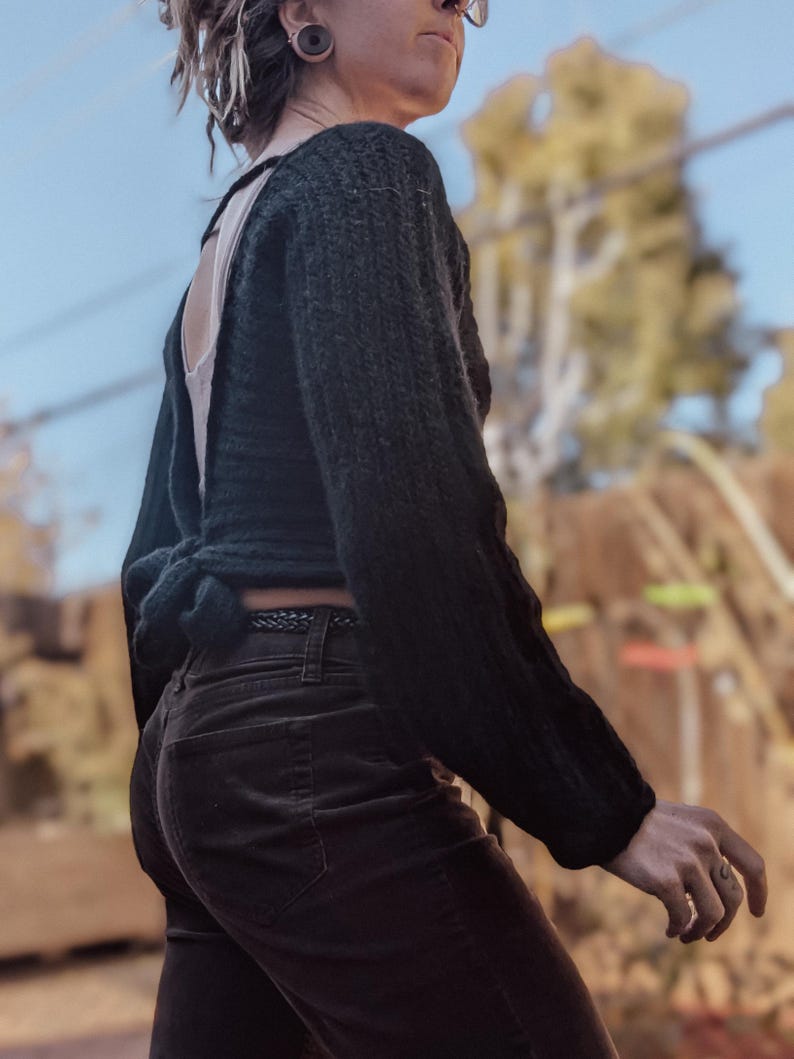 May include: A dark green, open-back sweater with long sleeves, tied at the waist. The sweater is paired with dark brown corduroy pants. The person is wearing glasses and has their hair styled in dreadlocks.