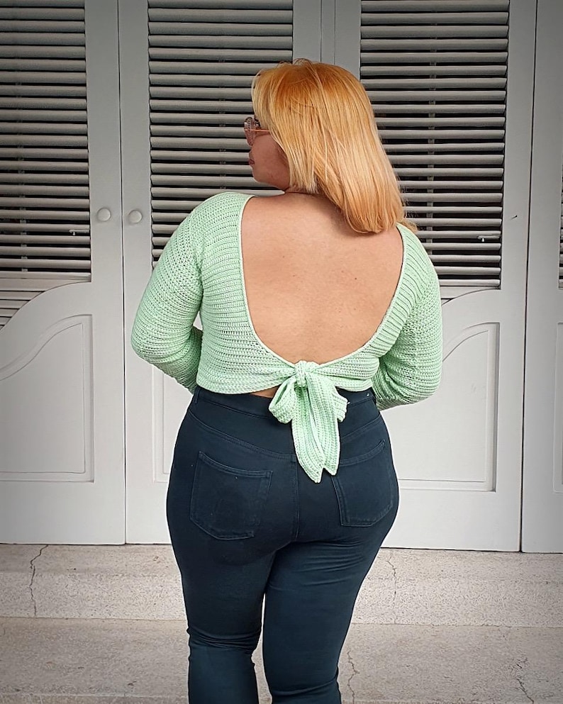 May include: A light green, open-back crochet top with long sleeves and a tie closure at the waist. The top is paired with dark blue jeans. The background features white doors with horizontal slats.