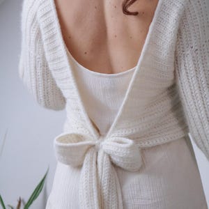 May include: Cream-colored, open-back sweater with long sleeves and a bow tie closure. The sweater is made of a chunky knit material and is paired with a cream-colored dress. The back of the sweater is open, revealing the wearer's back.
