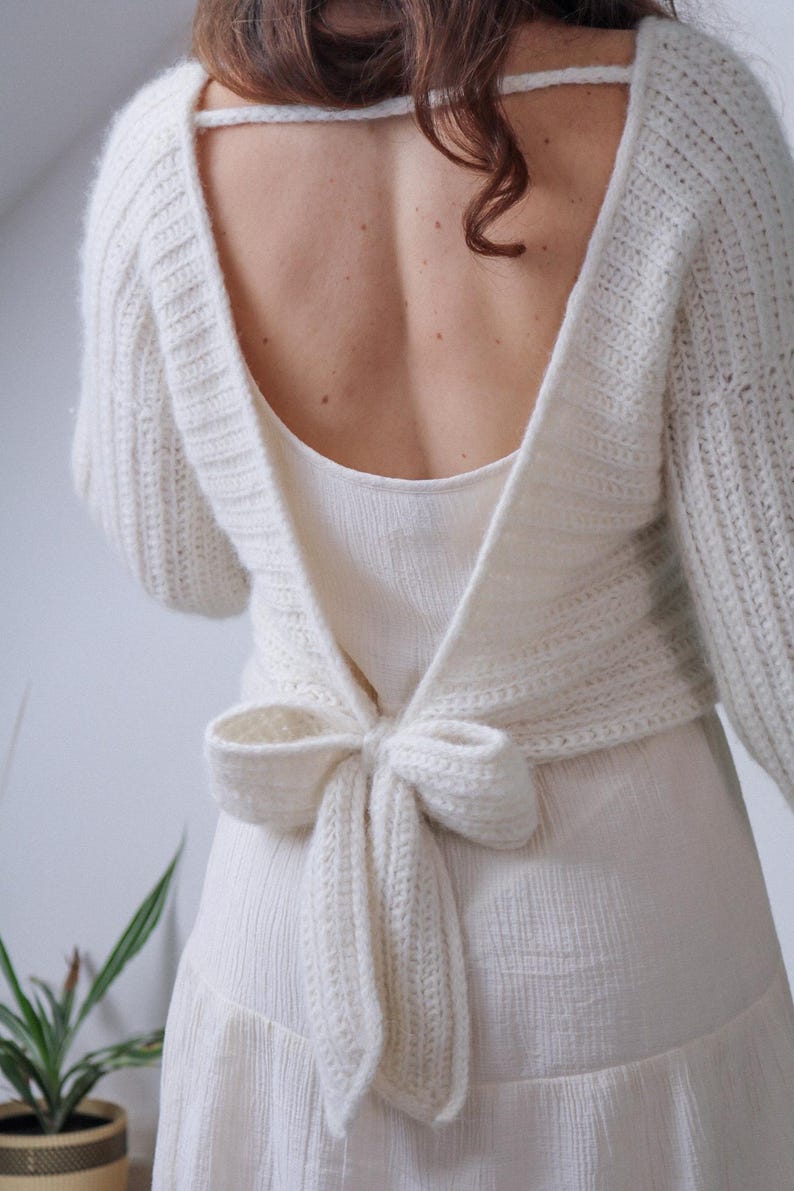 May include: Cream-colored, open-back sweater with long sleeves and a bow tie closure. The sweater is made of a chunky knit material and is paired with a cream-colored dress. The back of the sweater is open, revealing the wearer's back.