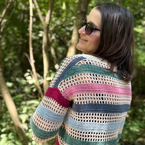 May include: A multi-colored, striped crochet sweater in shades of cream, green, blue, pink, and burgundy. The open-weave design is visible. The sweater has long sleeves and is worn outdoors.