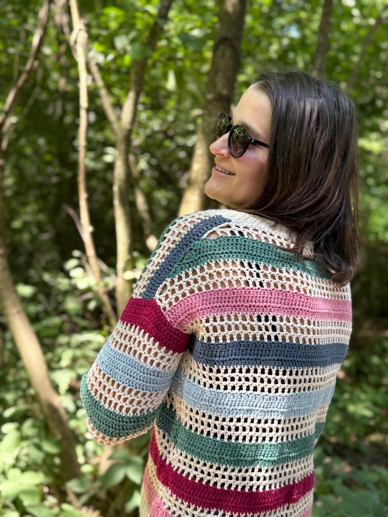 May include: A multi-colored, striped crochet sweater in shades of cream, green, blue, pink, and burgundy. The open-weave design is visible. The sweater has long sleeves and is worn outdoors.