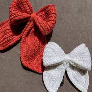 May include: Two crocheted bows, one red and one white, on a brown fabric background.