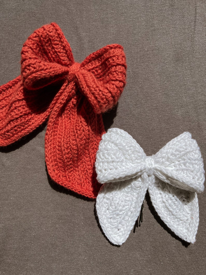 May include: Two crocheted bows, one red and one white, on a brown fabric background.