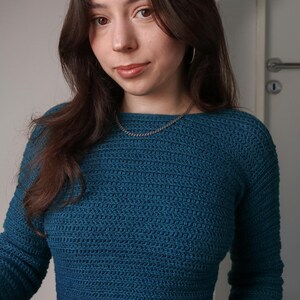 May include: Teal-colored crochet sweater with long sleeves. The sweater has a crew neck and a textured stitch pattern. A silver chain necklace is visible. The person is wearing black pants. The background is a neutral wall.