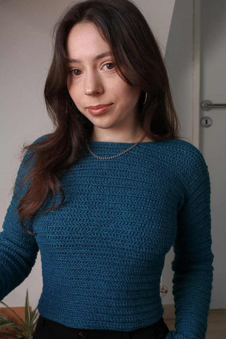 May include: Teal-colored crochet sweater with long sleeves. The sweater has a crew neck and a textured stitch pattern. A silver chain necklace is visible. The person is wearing black pants. The background is a neutral wall.