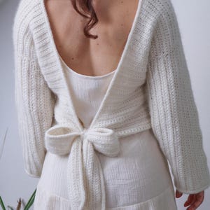 May include: Cream-colored, open-back sweater with long sleeves and a bow tie closure. The sweater is made of a chunky knit material. The model is wearing a white dress underneath.