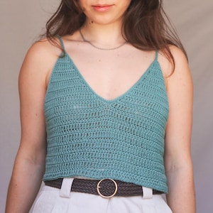 May include: A teal crochet top with a V-neck and spaghetti straps. The top is paired with white pants and a brown belt with a gold buckle. The top is a handmade garment.