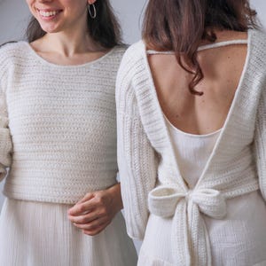 May include: Two people wearing off-white, hand-crocheted sweaters. One sweater has a boat neck and long sleeves with ribbed cuffs. The other has a low back and ties in a bow at the waist. Both are paired with white skirts.