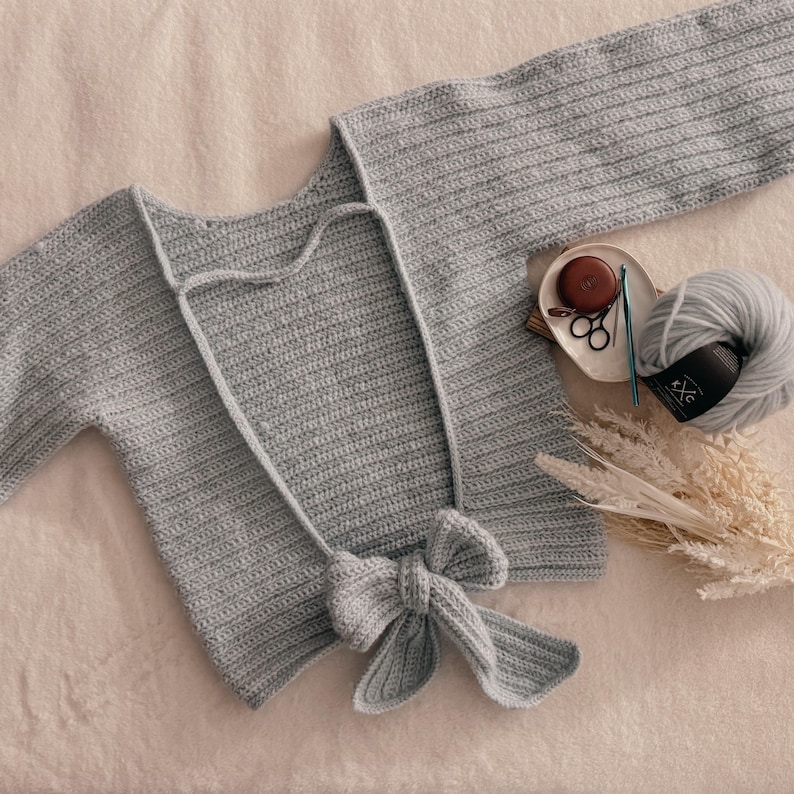 May include: A light gray, handmade crochet top with long sleeves and a bow detail at the waist. The top is displayed on a soft, neutral surface, with a ball of yarn, scissors, and a crochet hook nearby. The top is a delicate, handmade garment.