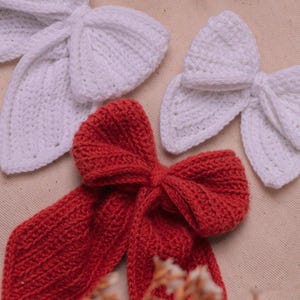 May include: Two white crocheted bows and one red crocheted bow. The bows are on a beige fabric background.
