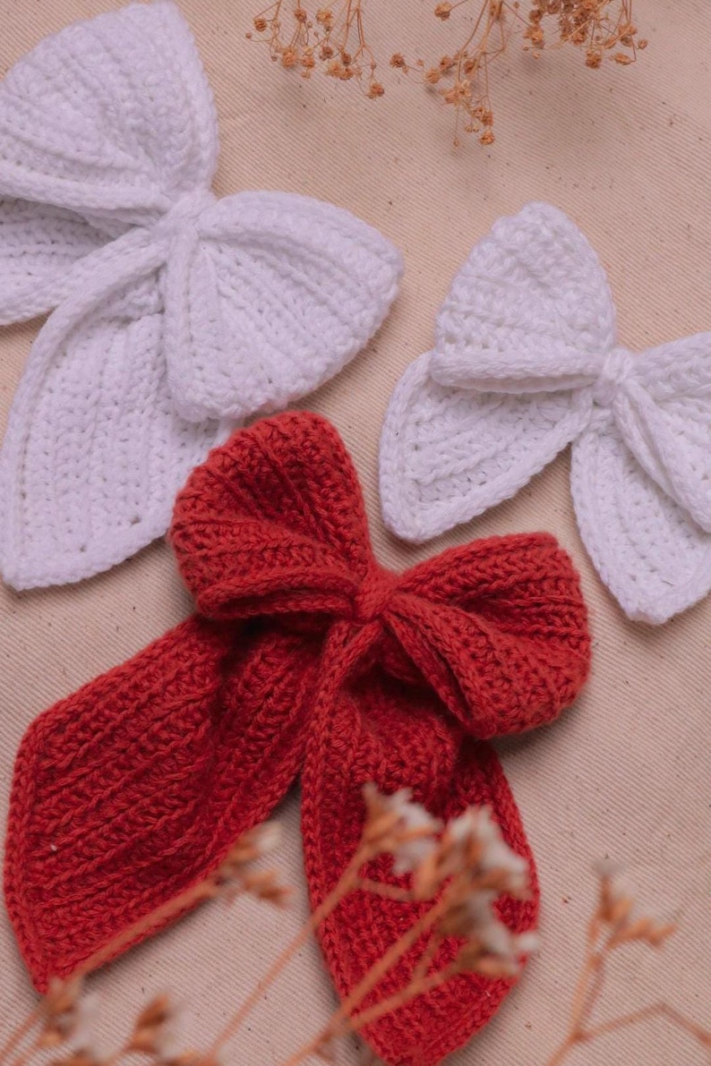May include: Two white crocheted bows and one red crocheted bow. The bows are on a beige fabric background.