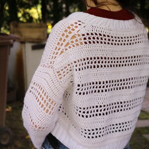 May include: A white, open-weave crochet sweater with a cropped fit. The sweater features a series of horizontal and vertical openwork patterns, creating a textured and airy design. The sleeves are long and the neckline is a crew neck.