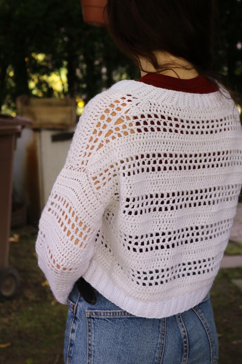 May include: A white, open-weave crochet sweater with a cropped fit. The sweater features a series of horizontal and vertical openwork patterns, creating a textured and airy design. The sleeves are long and the neckline is a crew neck.
