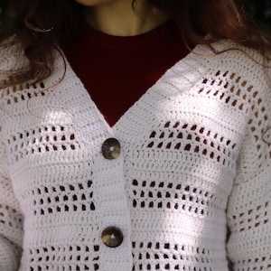 May include: A white, open-weave crochet cardigan with a V-neck and three large, round, brown buttons. The cardigan is worn over a dark red top. The crochet pattern features rectangular openings. The sleeves are long.