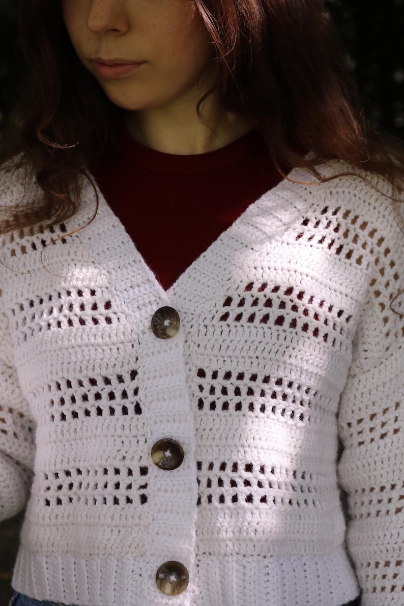 May include: A white, open-weave crochet cardigan with a V-neck and three large, round, brown buttons. The cardigan is worn over a dark red top. The crochet pattern features rectangular openings. The sleeves are long.
