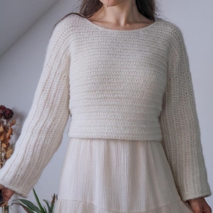 May include: Cream-colored crochet sweater with long sleeves and a scoop neckline. The sweater is paired with a light-colored, flowing skirt. The outfit is a casual, handmade style, suitable for everyday wear.