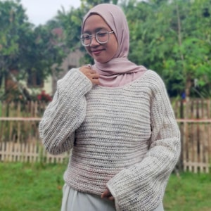 May include: A cream-colored, crocheted sweater with long sleeves. The sweater has a wide neckline and a textured, ribbed pattern. The sleeves are slightly flared. The sweater is paired with a light-colored skirt. The background features greenery and a wooden fence.