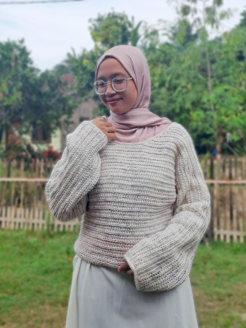 May include: A cream-colored, crocheted sweater with long sleeves. The sweater has a wide neckline and a textured, ribbed pattern. The sleeves are slightly flared. The sweater is paired with a light-colored skirt. The background features greenery and a wooden fence.
