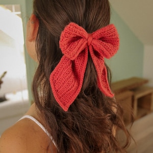 May include: A coral-coloured crocheted bow hair accessory. The bow is tied in a large, loose knot and has long, flowing tails.