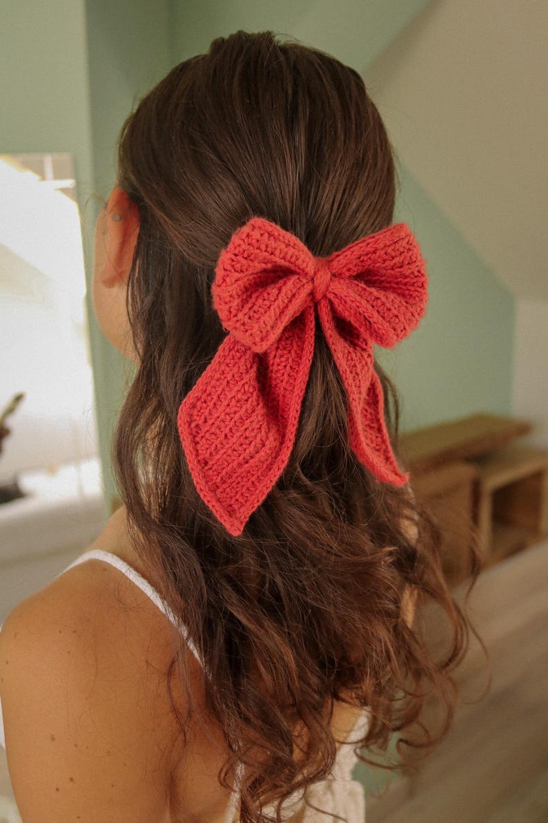 May include: A coral-coloured crocheted bow hair accessory. The bow is tied in a large, loose knot and has long, flowing tails.