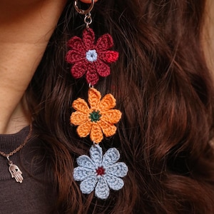 Può includere: Orecchini a fiori all'uncinetto, composti da tre fiori a forma di margherita nei colori bordeaux, arancione e azzurro. Ogni fiore ha un centro a contrasto. Stile bohémien fatto a mano, appesi a un cerchio d'argento.