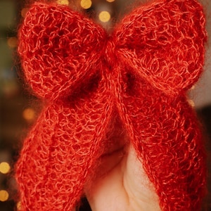 May include: A large, fluffy, red knitted bow. The bow is made of a soft, fuzzy yarn and has a loose, flowing shape.