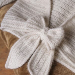 May include: Ivory-colored, knitted wrap with a bow closure. The wrap features a textured knit pattern and a bow detail at the front. The soft, off-white color and cozy texture suggest a warm, comfortable accessory.