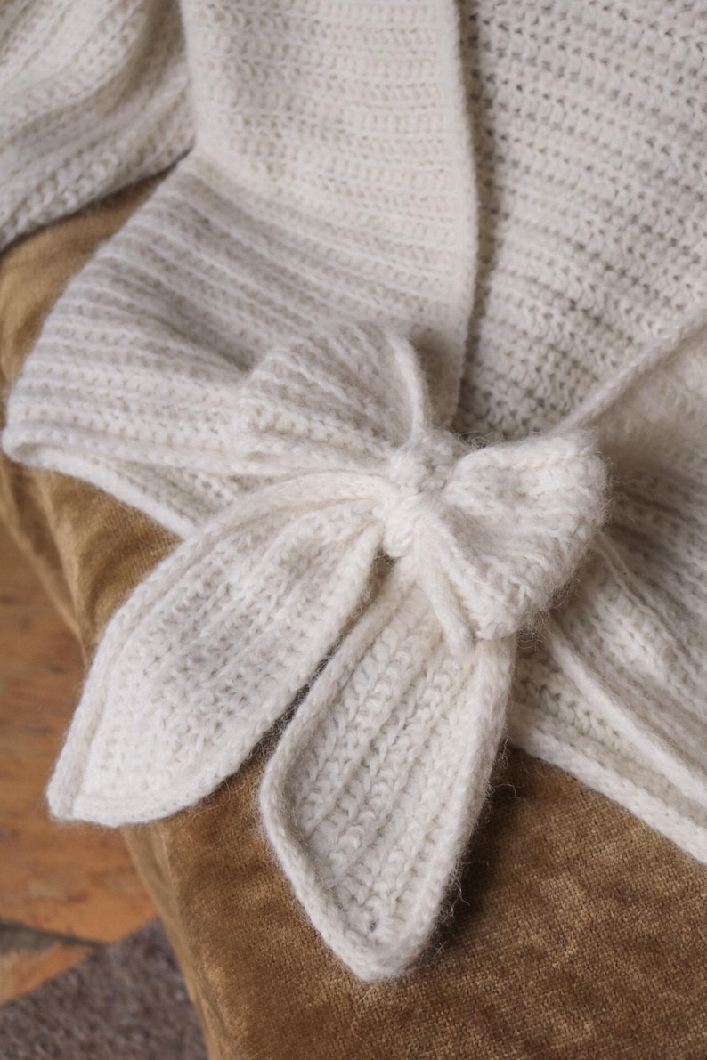 May include: Ivory-colored, knitted wrap with a bow closure. The wrap features a textured knit pattern and a bow detail at the front. The soft, off-white color and cozy texture suggest a warm, comfortable accessory.