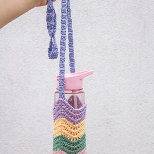 以下が含まれることがあります： 紫、ピンク、緑、黄色のストライプ模様のクロシェ編みの水筒ホルダー。ホルダーには長い紫色のストラップが付いています。