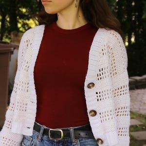 May include: A white, open-weave crochet cardigan with three brown buttons. The cardigan is worn over a maroon ribbed top and blue jeans with a black belt. The model is wearing silver hoop earrings.