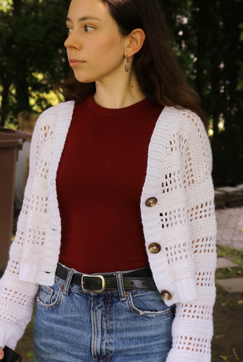 May include: A white, open-weave crochet cardigan with three brown buttons. The cardigan is worn over a maroon ribbed top and blue jeans with a black belt. The model is wearing silver hoop earrings.