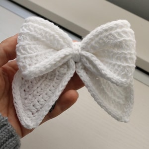 May include: A white crocheted bow with a textured design. The bow is tied in a knot in the centre.