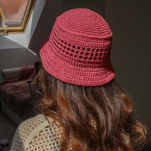 May include: A handmade, rose-colored crochet bucket hat. The hat features a textured, openwork design around the crown and a solid brim. The hat is shown from the back, with long, wavy brown hair visible.