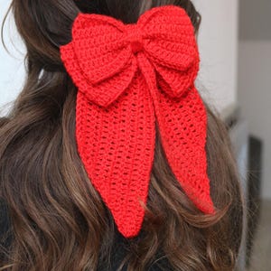 May include: A vibrant red, crocheted hair bow. The bow is large and has a classic bow shape with long, flowing tails. The hair accessory is set against a backdrop of brown hair.