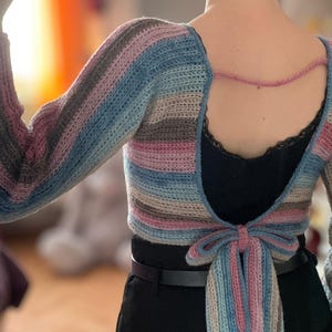 May include: A handmade, open-back crochet top with long sleeves. The top features horizontal stripes in shades of blue, pink, gray, and beige. A pink cord connects the shoulders, and a bow detail is at the waist. The top is worn over a black garment.