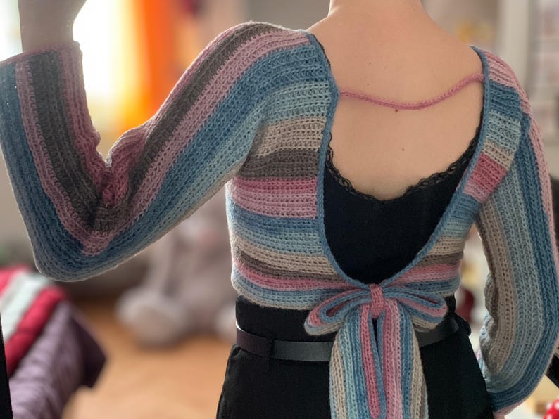 May include: A handmade, open-back crochet top with long sleeves. The top features horizontal stripes in shades of blue, pink, gray, and beige. A pink cord connects the shoulders, and a bow detail is at the waist. The top is worn over a black garment.