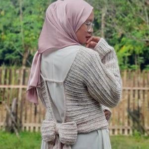 May include: A light beige, open-back sweater with a large bow detail at the waist. The sweater has long sleeves and a ribbed texture. The person is wearing a light pink headscarf and glasses. The background is a blurred view of greenery and a wooden fence.