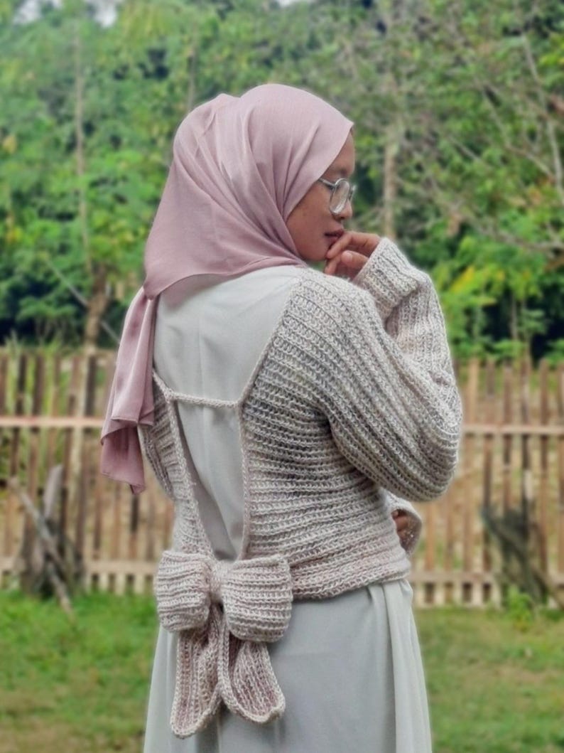 May include: A light beige, open-back sweater with a large bow detail at the waist. The sweater has long sleeves and a ribbed texture. The person is wearing a light pink headscarf and glasses. The background is a blurred view of greenery and a wooden fence.