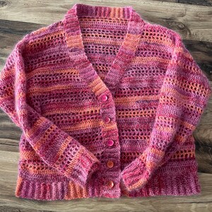 May include: A handmade, open-weave cardigan in shades of pink and orange. The button-down sweater features a V-neck and long sleeves. The crochet design has a textured appearance, with a mix of solid and open stitch patterns.
