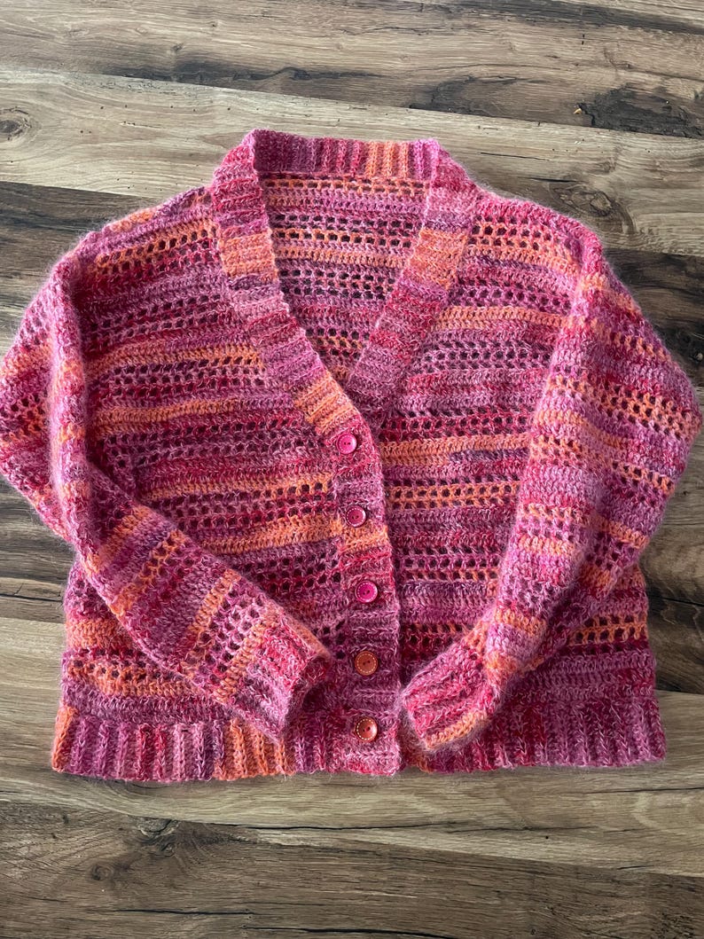 May include: A handmade, open-weave cardigan in shades of pink and orange. The button-down sweater features a V-neck and long sleeves. The crochet design has a textured appearance, with a mix of solid and open stitch patterns.